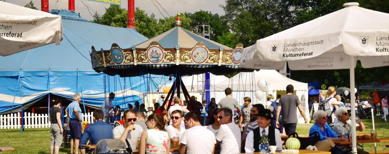 Menschen sitzen an Holztischen unter Regenschirmen auf dem Festplatz mit Zirkuszelt und Karussell im Hintergrund. Es ist ein sonniger Tag bei den Stadtteilwochen München!