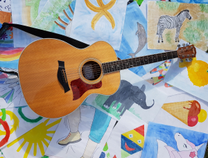 Eine hölzerne Akustikgitarre liegt auf farbenfrohen Kinderzeichnungen, die ein Zebra, einen Elefanten, einen Delfin, einen Regenbogen, eine Sonne, eine Eistüte und andere spielerische Szenen zeigen.