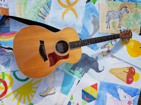Eine hölzerne Akustikgitarre liegt auf farbenfrohen Kinderzeichnungen, die ein Zebra, einen Elefanten, einen Delfin, einen Regenbogen, eine Sonne, eine Eistüte und andere spielerische Szenen zeigen.
