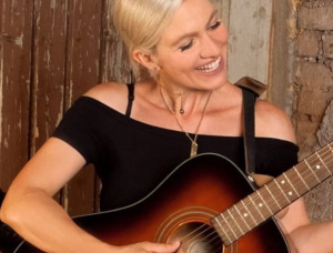 Eine lächelnde Frau mit kurzen blonden Haaren spielt eine akustische Gitarre. Sie trägt ein schwarzes schulterfreies Oberteil und eine Halskette und steht vor einem rustikalen Hintergrund aus Holz und Ziegeln.