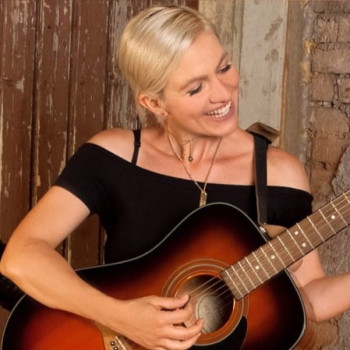 Eine lächelnde Frau mit kurzen blonden Haaren spielt eine akustische Gitarre. Sie trägt ein schwarzes schulterfreies Oberteil und eine Halskette und steht vor einem rustikalen Hintergrund aus Holz und Ziegeln.
