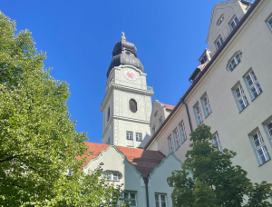 Ein hohes, helles Gebäude mit einem Uhrenturm und einem markanten Kuppeldach erhebt sich über roten Dächern und grünen Bäumen vor einem klaren blauen Himmel.