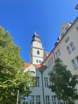 Ein hohes, helles Gebäude mit einem Uhrenturm und einem markanten Kuppeldach erhebt sich über roten Dächern und grünen Bäumen vor einem klaren blauen Himmel.