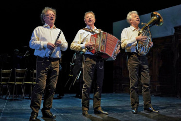Drei ältere Männer stehen in passenden weißen Hemden und dunklen Hosen auf der Bühne und spielen jeweils ein Instrument: Klarinette, Akkordeon und Horn. Sie spielen im Scheinwerferlicht auf der Bühne.