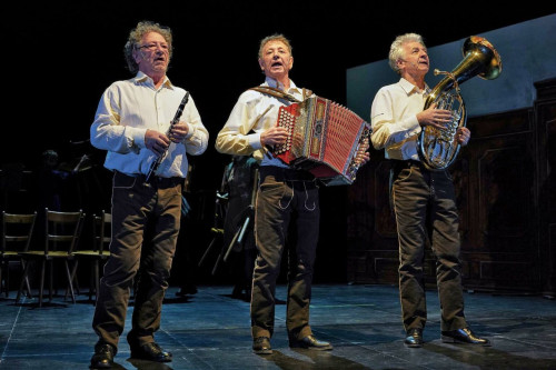 Drei ältere Männer stehen in passenden weißen Hemden und dunklen Hosen auf der Bühne und spielen jeweils ein Instrument: Klarinette, Akkordeon und Horn. Sie spielen im Scheinwerferlicht auf der Bühne.
