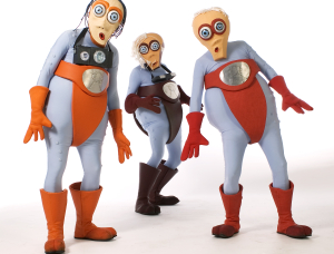 Drei Personen in cartoonhaften Kostümen mit Käferaugen, orangefarbenen Handschuhen, Stiefeln und roten oder braunen Slips posieren vor einem weißen Hintergrund. Alle haben einen übertriebenen Gesichtsausdruck und große, runde Silbermedaillons auf der Brust.