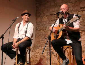 Zwei Männer sitzen auf Hockern auf der Bühne. Der eine, mit Hut und Hosenträgern, lächelt, während der andere, in eine Weste gekleidet, in ein Mikrofon singt und eine akustische Gitarre spielt. Im Hintergrund ist eine Steinmauer zu sehen.