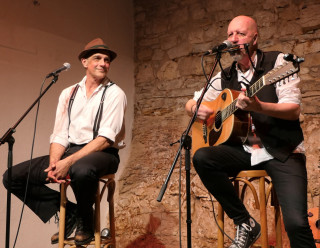 Zwei Männer sitzen auf Hockern auf der Bühne. Der eine, mit Hut und Hosenträgern, lächelt, während der andere, in eine Weste gekleidet, in ein Mikrofon singt und eine akustische Gitarre spielt. Im Hintergrund ist eine Steinmauer zu sehen.
