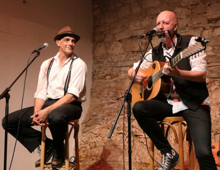 Zwei Männer sitzen auf Hockern auf der Bühne. Der eine, mit Hut und Hosenträgern, lächelt, während der andere, in eine Weste gekleidet, in ein Mikrofon singt und eine akustische Gitarre spielt. Im Hintergrund ist eine Steinmauer zu sehen.