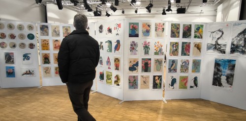 Eine Person in einer schwarzen Jacke geht in einem gut beleuchteten Kunstausstellungsraum an mehreren Schautafeln mit farbenfrohen Gemälden und Zeichnungen vorbei, die hauptsächlich Tiere, Blumen und Natur darstellen.