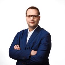 Alexander Friedich in dunkelblauem Anzug steht mit verschränkten Armen und blickt selbstbewusst in die Kamera vor einem schlichten weißen Hintergrund.