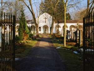 Das Bild zeigt den Eingang zu einem Friedhof mit einem offenen Eisentor, einem von Bäumen gesäumten Weg, Grabsteinen auf beiden Seiten und einer weißen Kapelle mit einem Kreuz im Hintergrund bei klarem Tageslicht.
