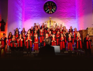 Ein großer Chor in schwarzen Kostümen und mit roten Schals singt auf der Bühne einer Kirche, die von farbenfrohem lila und rosa Licht erhellt wird. Das Publikum schaut zu, und ein Dirigent steht vor dem Chor und blickt ihm zu.