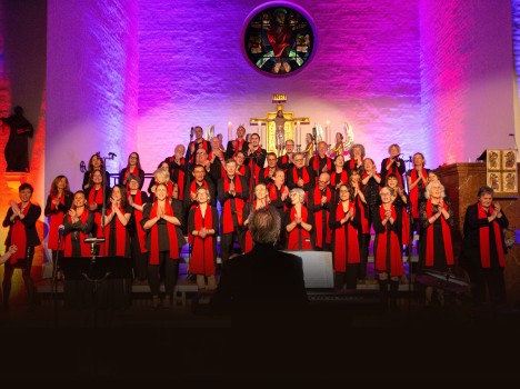 Ein großer Chor in schwarzen Kostümen und mit roten Schals singt auf der Bühne einer Kirche, die von farbenfrohem lila und rosa Licht erhellt wird. Das Publikum schaut zu, und ein Dirigent steht vor dem Chor und blickt ihm zu.