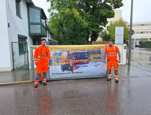 Zwei Männer vom Straßenunterhaltungsbezirk Süd in leuchtend orangefarbenen Arbeitsuniformen stehen an einer Einfahrt und halten ein großes Transparent mit dem Bild eines Winterdienstfahrzeugs und einem deutschen Text über die Vorbereitung auf das Winterwetter. Im Hintergrund sind Häuser und Bäume zu sehen.