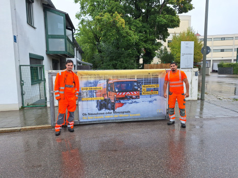 Zwei Männer vom Straßenunterhaltungsbezirk Süd in leuchtend orangefarbenen Arbeitsuniformen stehen an einer Einfahrt und halten ein großes Transparent mit dem Bild eines Winterdienstfahrzeugs und einem deutschen Text über die Vorbereitung auf das Winterwetter. Im Hintergrund sind Häuser und Bäume zu sehen.