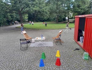 Zwei Holzstühle und ein Tisch mit Getränken und Snacks stehen auf einem gepflasterten Platz in der Nähe eines roten Schuppens. Im Vordergrund stehen bunte Tüten und Flaschen; Menschen spielen und gehen in einem grasbewachsenen Park mit Bäumen im Hintergrund.
