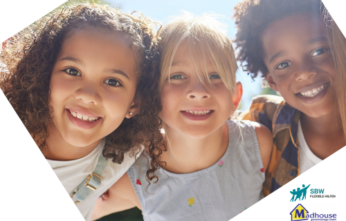 Drei lächelnde Kinder stehen dicht beieinander im Freien im Sonnenlicht. Zwei Mädchen, eines mit lockigem und eines mit hellblondem Haar, und ein Junge mit lockigem Haar schauen fröhlich in die Kamera. In den Ecken sind SBW- und Madhouse-Logos zu sehen.