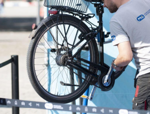 Eine Person in einem hellgrauen Hemd repariert das Hinterrad eines schwarzen Fahrrads, das in einem Fahrradständer angehoben ist. Das Gesicht der Person ist nicht zu erkennen. Der Hintergrund ist unscharf.