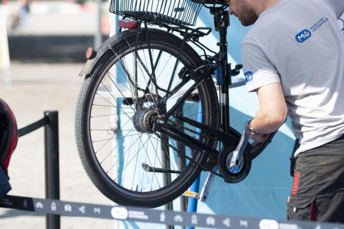 Eine Person in einem hellgrauen Hemd repariert das Hinterrad eines schwarzen Fahrrads, das in einem Fahrradständer angehoben ist. Das Gesicht der Person ist nicht zu erkennen. Der Hintergrund ist unscharf.