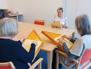 Drei Frauen sitzen um einen Tisch herum, zwei spielen Holzsaiteninstrumente und lesen dabei Noten, und eine leitet die Sitzung mit einem Notenblatt vor sich.