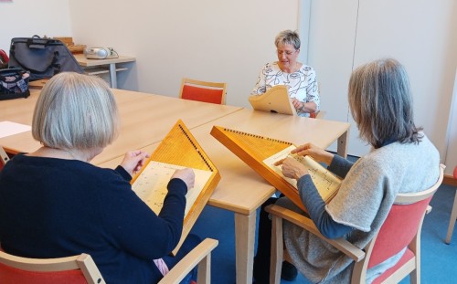 Drei Frauen sitzen um einen Tisch herum, zwei spielen Holzsaiteninstrumente und lesen dabei Noten, und eine leitet die Sitzung mit einem Notenblatt vor sich.
