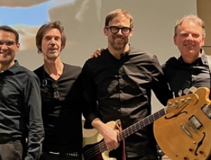 Vier Männer in dunkler Kleidung stehen lächelnd zusammen. Einer hält eine Gitarre, ein anderer einen E-Bass. Sie posieren vor einem hellen Hintergrund.