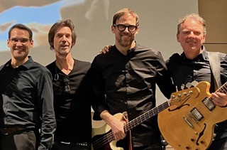 Vier Männer in dunkler Kleidung stehen lächelnd zusammen. Einer hält eine Gitarre, ein anderer einen E-Bass. Sie posieren vor einem hellen Hintergrund.