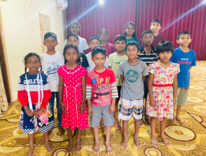Eine Gruppe von fünfzehn Kindern steht in Reihen auf einem gemusterten Teppich, barfuß und mit dem Gesicht zur Kamera. Im Hintergrund ist ein roter Vorhang zu sehen, und die Kinder sind in bunter Freizeitkleidung gekleidet.