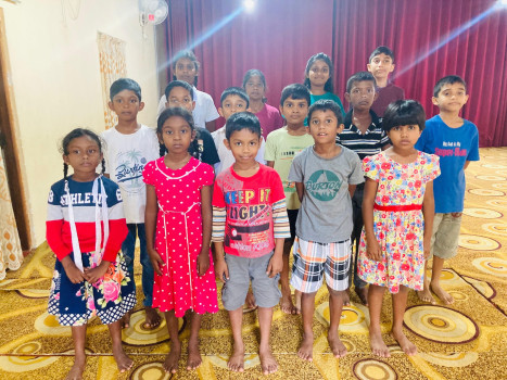 Eine Gruppe von fünfzehn Kindern steht in Reihen auf einem gemusterten Teppich, barfuß und mit dem Gesicht zur Kamera. Im Hintergrund ist ein roter Vorhang zu sehen, und die Kinder sind in bunter Freizeitkleidung gekleidet.