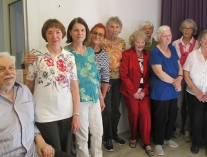 Eine Gruppe von elf älteren Erwachsenen, zehn Frauen und ein Mann, steht zusammen im Haus und lächelt in die Kamera. Sie tragen Freizeitkleidung und stehen neben einem Fenster mit hellen Vorhängen.