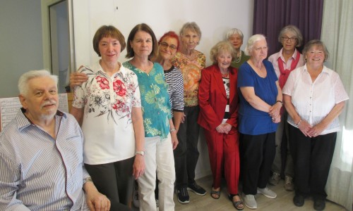 Eine Gruppe von elf älteren Erwachsenen, zehn Frauen und ein Mann, steht zusammen im Haus und lächelt in die Kamera. Sie tragen Freizeitkleidung und stehen neben einem Fenster mit hellen Vorhängen.