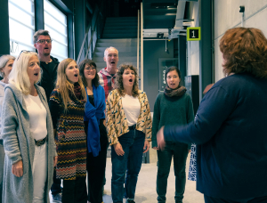 Eine Gruppe von acht Personen steht in einem Halbkreis und singt gemeinsam, während ein Dirigent sie in ein modernes Gebäude mit Treppen und großen Fenstern dirigiert.