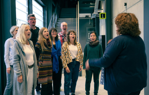 Eine Gruppe von acht Personen steht in einem Halbkreis und singt gemeinsam, während ein Dirigent sie in ein modernes Gebäude mit Treppen und großen Fenstern dirigiert.