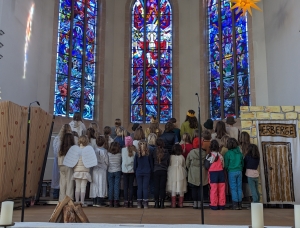 Eine Gruppe kostümierter Kinder, darunter einige mit Engelsflügeln, steht mit dem Rücken zur Kamera vor bunten Buntglasfenstern in einer Kirche. Rechts befindet sich eine Tür mit der Aufschrift „Herberge“.