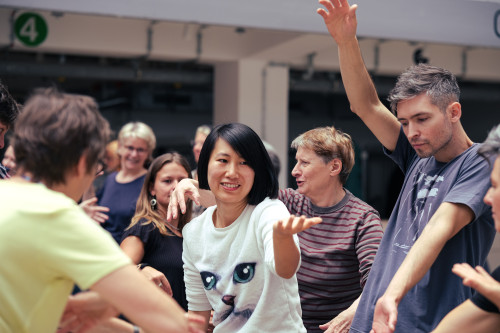 Eine Gruppe von Menschen unterschiedlichen Alters steht in einem Innenraum dicht beieinander, lächelt und tanzt mit erhobenen Armen. Die Stimmung ist energisch und fröhlich, und die Frau in der Mitte trägt ein weißes Hemd mit einem großen Katzengesicht als Aufdruck.