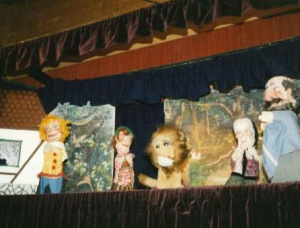Ein Puppenspiel mit fünf bunten Puppen, darunter ein Löwe und vier menschliche Figuren, wird auf einer Bühne mit dunklen Vorhängen und gemalten Waldkulissen aufgeführt.