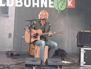 Ein Mann mit grauen Haaren und Bart spielt eine Akustikgitarre und singt in ein Mikrofon auf einer Freilichtbühne. Er sitzt, und um ihn herum befinden sich Audiogeräte und Kabel. Im Hintergrund ist ein Teil eines Veranstaltungsbanners zu sehen.