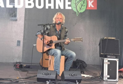Ein Mann mit grauen Haaren und Bart spielt eine Akustikgitarre und singt in ein Mikrofon auf einer Freilichtbühne. Er sitzt, und um ihn herum befinden sich Audiogeräte und Kabel. Im Hintergrund ist ein Teil eines Veranstaltungsbanners zu sehen.