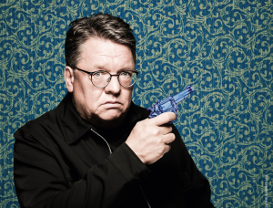 Ein ernst dreinblickender Mann mit Brille und schwarzer Jacke posiert vor einem gemusterten blauen und grünen Hintergrund und hält eine blaue Spielzeugpistole in der Hand.