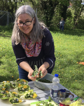 Eine lächelnde Frau mit grauem Haar und Brille steht draußen an einem Tisch und hält Grünzeug in der Hand. Auf dem Tisch stehen Teller mit Grünzeug und Blumen, eine Schale und Wasserflaschen. Im Hintergrund sind Bäume und Gras zu sehen, hinter denen eine weitere Person geht.