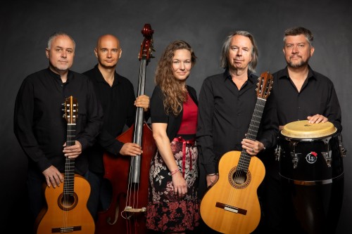 Fünf Musiker posieren vor einem dunklen Hintergrund und halten Instrumente wie zwei Gitarren, einen Kontrabass und Conga-Trommeln in den Händen. Die Gruppe, in schwarz-roter Kleidung, steht dicht beieinander und blickt in die Kamera.