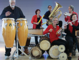 Sechs Erwachsene spielen verschiedene Musikinstrumente, darunter Conga-Trommeln, Marimba, Tuba, Posaune, Flöte und Handtrommeln, in einem hell erleuchteten Raum mit einem hellblauen und lila Hintergrund. Alle lächeln und scheinen sich zu amüsieren.