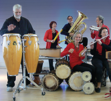 Sechs Erwachsene spielen verschiedene Musikinstrumente, darunter Conga-Trommeln, Marimba, Tuba, Posaune, Flöte und Handtrommeln, in einem hell erleuchteten Raum mit einem hellblauen und lila Hintergrund. Alle lächeln und scheinen sich zu amüsieren.