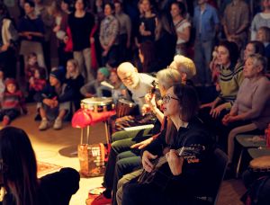 Eine Gruppe von Menschen versammelt sich in einem Raum, einige sitzend, andere stehend, und hört Live-Musik. Mehrere Personen spielen Instrumente wie Gitarre und Schlagzeug. Das Publikum scheint engagiert und aufmerksam zu sein.