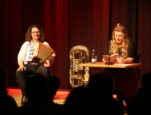 Ein Mann spielt Akkordeon neben einer großen Messingtuba, während eine Frau an einem Tisch sitzt und in ein Mikrofon aus einem Buch liest. Beide stehen auf der Bühne vor Publikum, hinter ihnen ein roter Bühnenvorhang.
