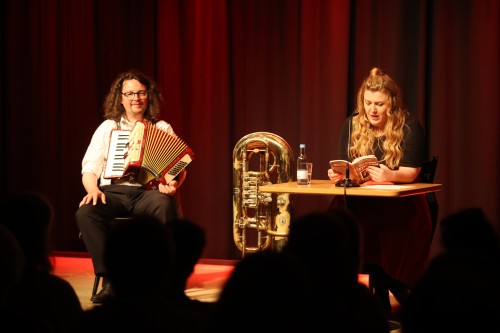 Ein Mann spielt Akkordeon neben einer großen Messingtuba, während eine Frau an einem Tisch sitzt und in ein Mikrofon aus einem Buch liest. Beide stehen auf der Bühne vor Publikum, hinter ihnen ein roter Bühnenvorhang.