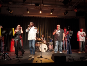 Auf der Bühne tritt eine Band mit sechs stehenden Personen und einem Schlagzeuger auf, der an einem Schlagzeug sitzt. Sie singen und spielen Instrumente unter warmem Bühnenlicht, hinter ihnen ein Vorhang.