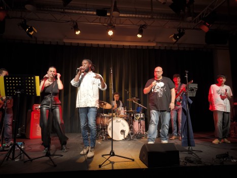 Auf der Bühne tritt eine Band mit sechs stehenden Personen und einem Schlagzeuger auf, der an einem Schlagzeug sitzt. Sie singen und spielen Instrumente unter warmem Bühnenlicht, hinter ihnen ein Vorhang.