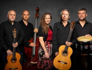 Fünf Musiker posieren vor einem dunklen Hintergrund und halten Instrumente wie zwei Gitarren, einen Kontrabass und Conga-Trommeln in den Händen. Die Gruppe, in schwarz-roter Kleidung, steht dicht beieinander und blickt in die Kamera.